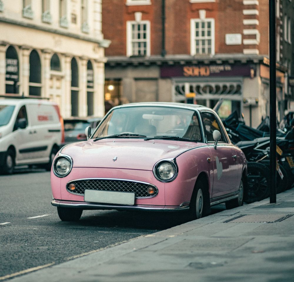 Baby pink Nissan Figaro parked at the side of the road in a city or town