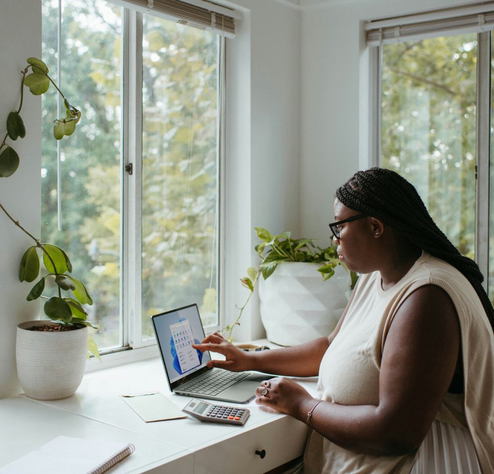 Woman working on a laptop at a desk near windows