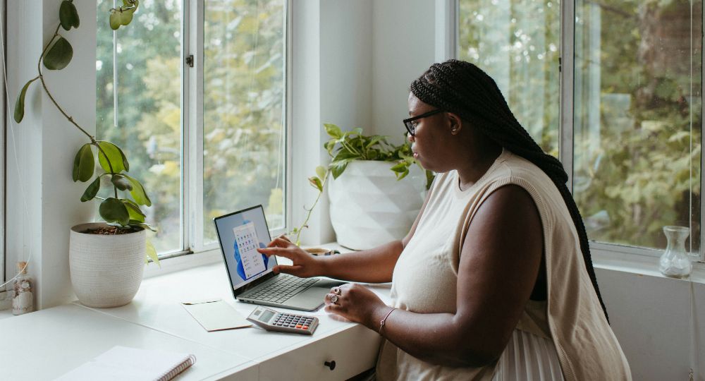 Woman working on a laptop at a desk near windows