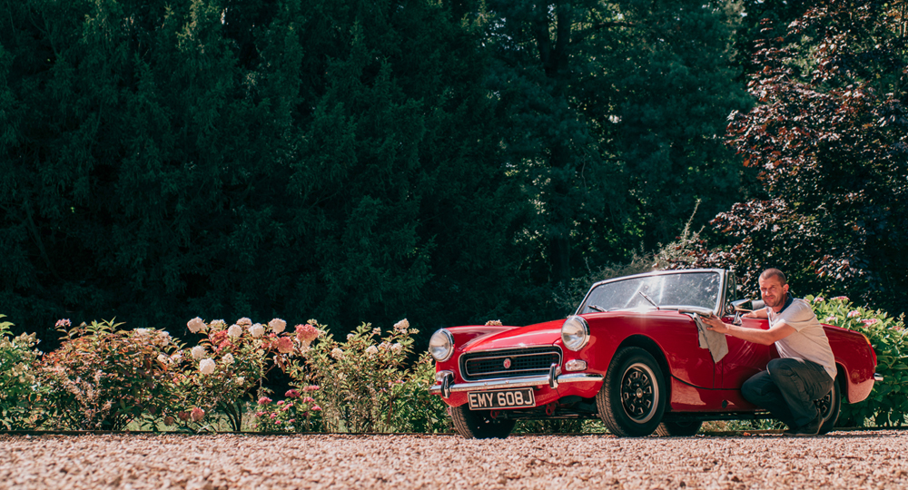 Man wiping down red classic convertible on a summer's day with trees in the background