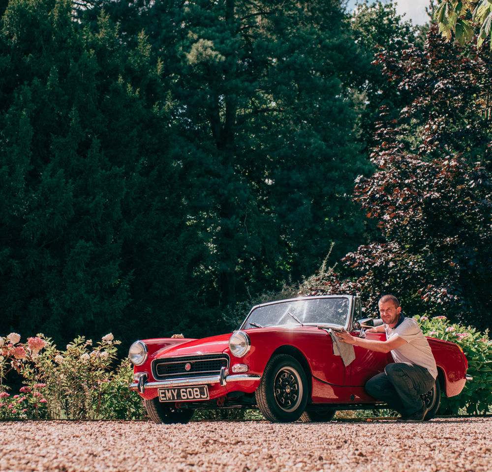 Man wiping down red classic convertible on a summer's day with trees in the background