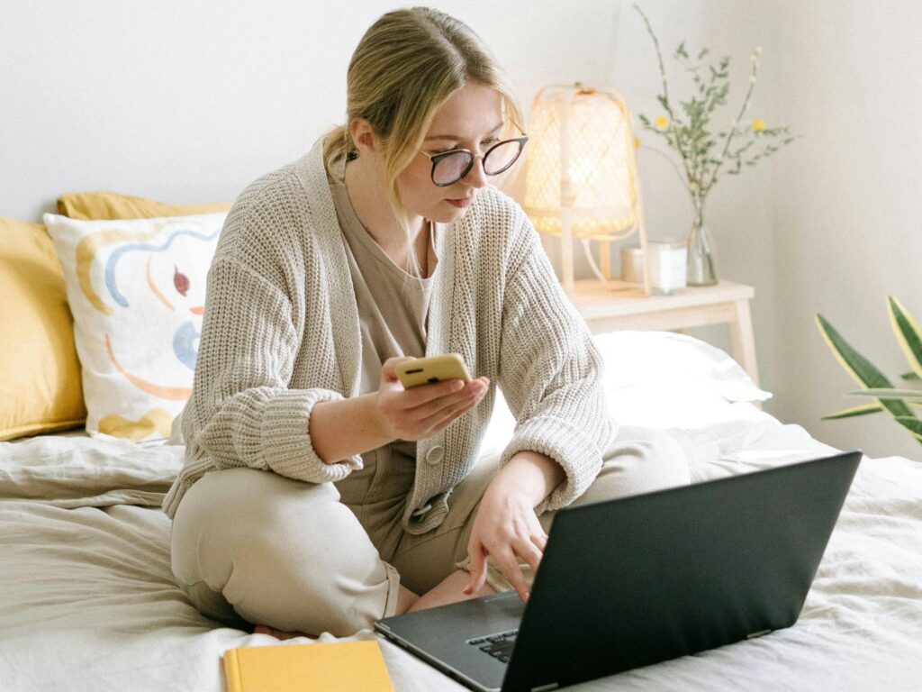 Woman sat crossed legged on bed, using laptop and smartphone - doing research