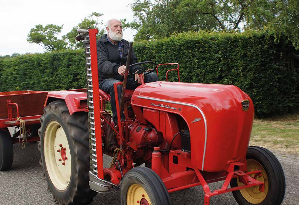 Man driving a classic red tractor down a road with green bushes and trees in the background