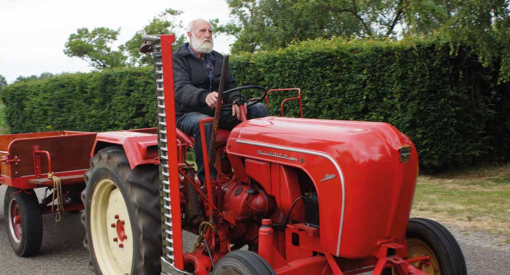 Man driving a classic red tractor down a road with green bushes and trees in the background