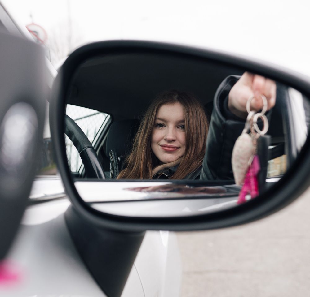 Side mirror image of a young driver at the wheel holding her car keys