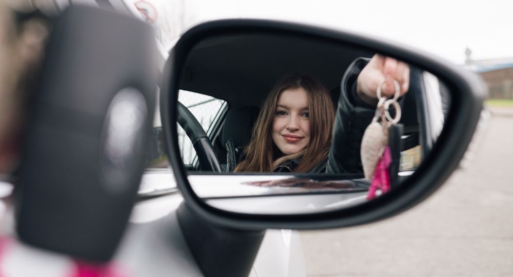 Side mirror image of a young driver at the wheel holding her car keys